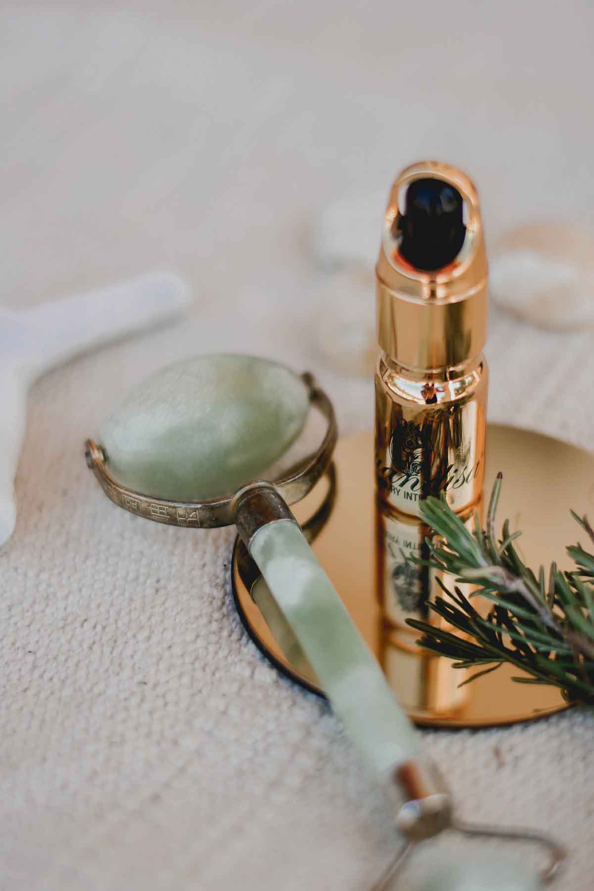 A jade facial roller and a gold-capped skincare serum bottle rest on a round mirror, with a sprig of greenery nearby, all set on a textured surface.