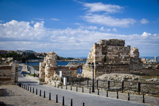 Ancient stone ruins stand near a cobblestone path by the water, with a blue sky and scattered clouds overhead. A black iron fence lines the walkway, and distant buildings are visible across the bay.