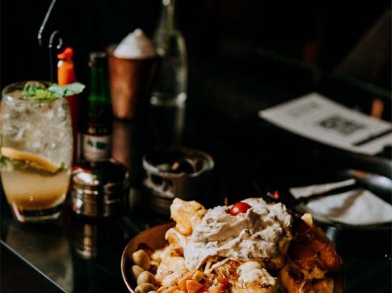 A close-up of a plate with nachos topped with whipped cream and a cherry, next to a cocktail with mint and lemon on a reflective table with condiments and utensils blurred in the background.