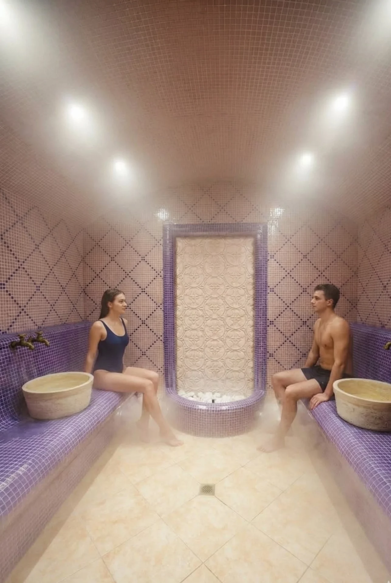 Ein gekacheltes Hoteldampfbad mit geschwungenen Wänden und Decken, violetten und rosafarbenen Mosaikbänken, zwei Steinwaschbecken und einem Zierbrunnen in der Mitte der Rückwand. Helle Lichter erhellen den einladenden Raum.